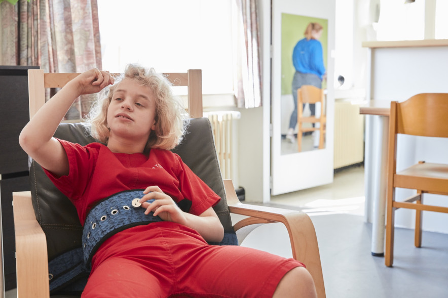 Blond meisje met beperking in rode kledij zit in zetelstoel in leefruimte Blond meisje met beperking in rode kledij zit in zetelstoel in leefruimte